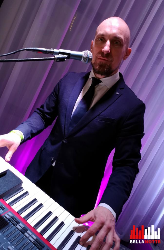 Muzyk w ciemnym garniturze, grający na keyboardzie i śpiewający do mikrofonu na tle fioletowych zasłon, z widocznym logo zespołu w prawym dolnym rogu.