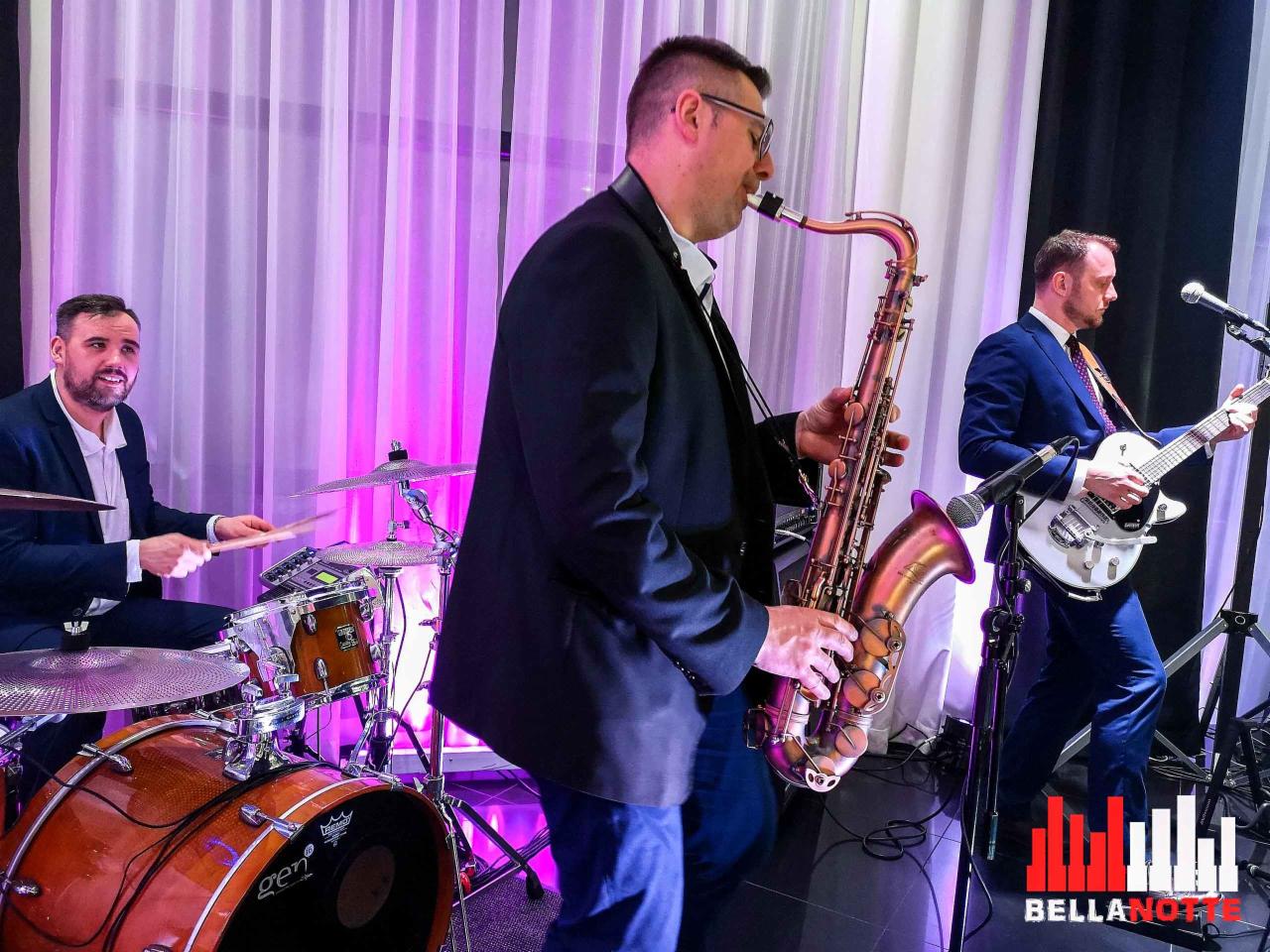 Trzech muzyków w trakcie występu: saksofonista w garniturze z saksofonem tenorowym, gitarzysta w niebieskim garniturze z białą gitarą elektryczną oraz perkusista z uśmiechem grający na perkusji.