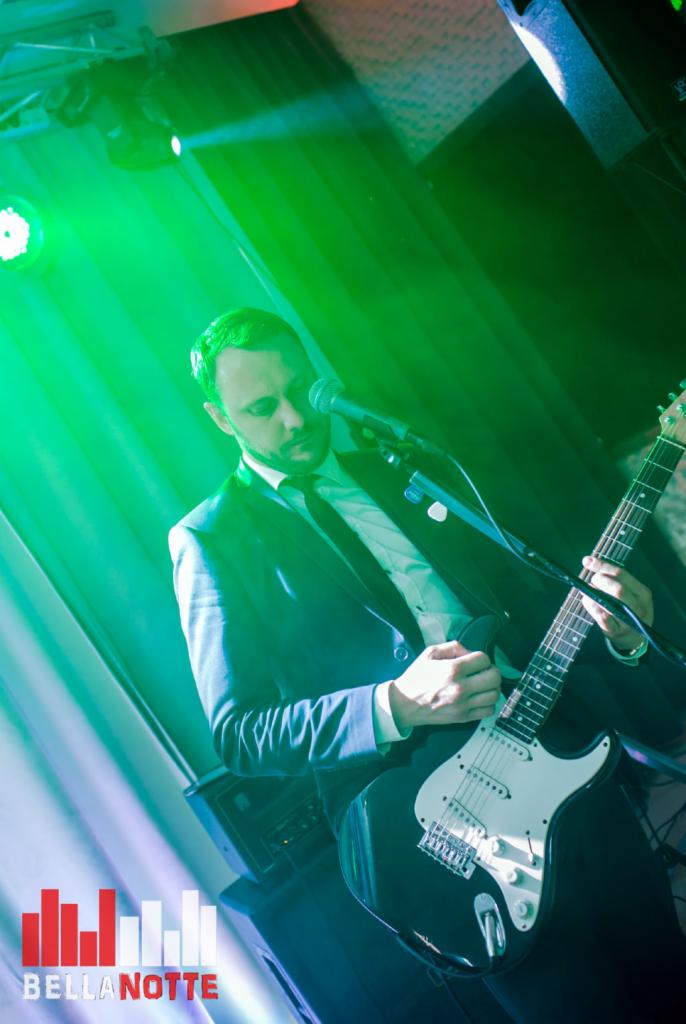 Mężczyzna w garniturze gra na czarnej gitarze elektrycznej pod zielonym oświetleniem scenicznym, widoczny mikrofon i logo zespołu BellaNotte w lewym dolnym rogu.