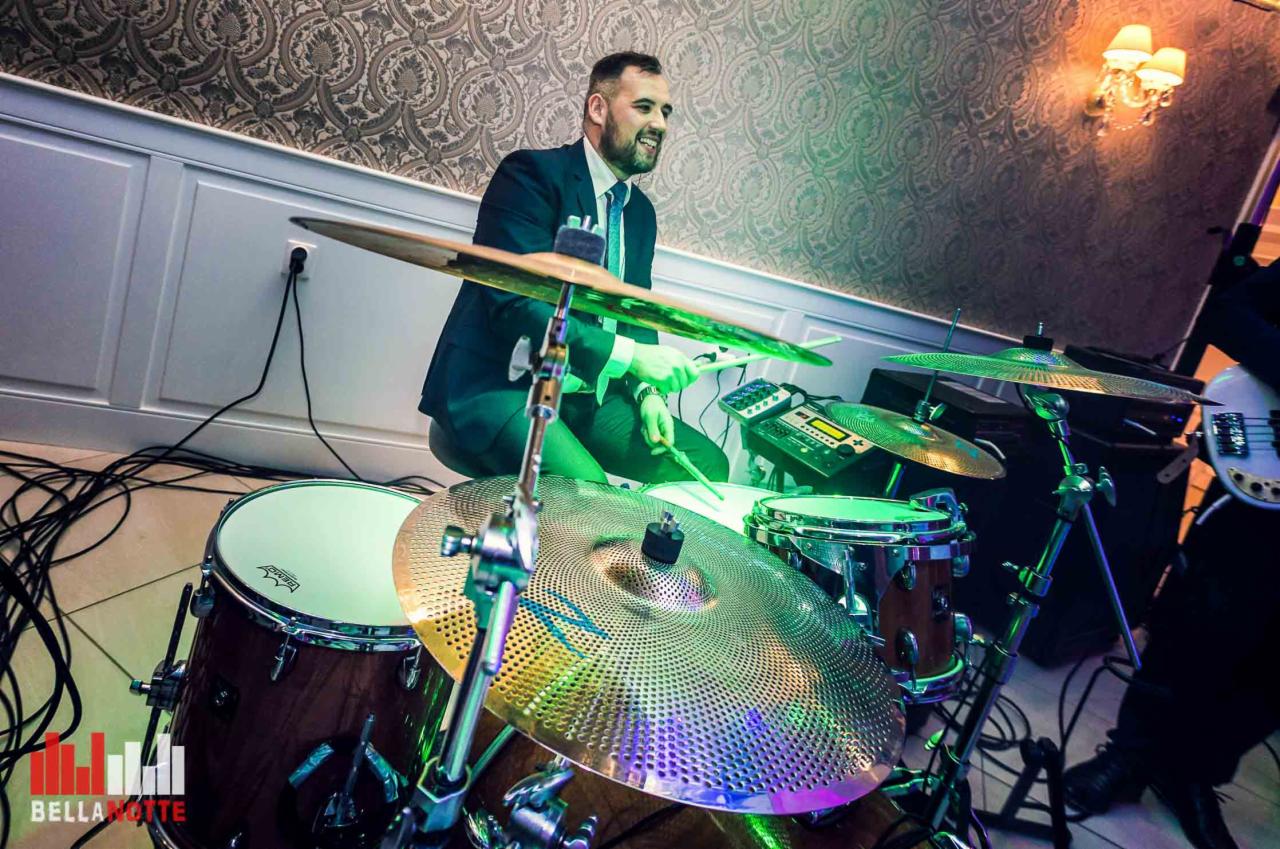 Perkusista w garniturze gra na perkusji podświetlonej zielonym światłem, widoczny fragment gitary basowej, na ścianie wzorzysta tapeta i kinkiet.