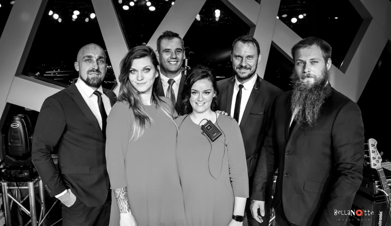 Czarno-białe zdjęcie zespołu muzycznego Bella Notte w garniturach i sukienkach, z gitarą basową widoczną po prawej stronie, na tle sceny z reflektorami.