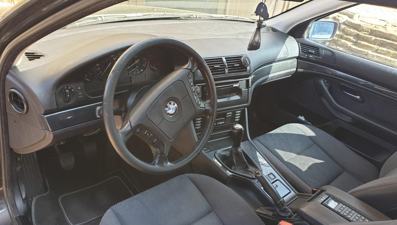 Wnętrze starszego modelu BMW serii 5 E39, widok na kierownicę z logo, deskę rozdzielczą, fotele z szarej tkaniny i czarną gałkę zmiany biegów; na lusterku wisi zapach samochodowy.