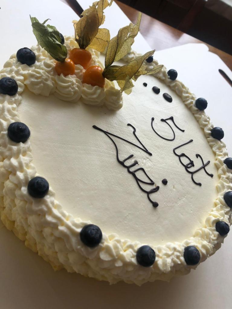Okrągły tort urodzinowy z białym kremem, dekorowany świeżymi borówkami, miechunką peruwiańską i napisem wykonanym ciemną polewą 'Na 5 lat'.