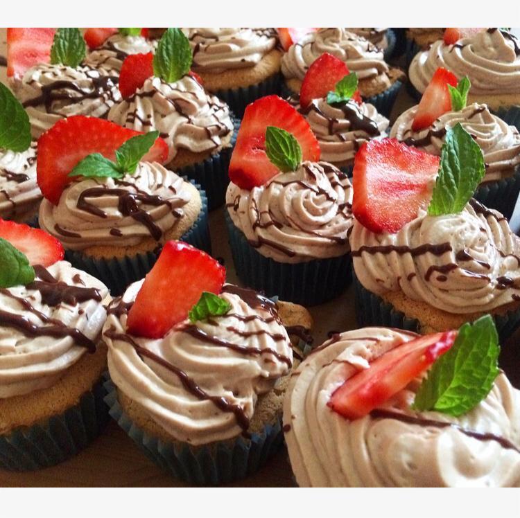 Rząd babeczek z kremem kawowym, polewą czekoladową, plasterkiem truskawki i listkiem mięty, w niebieskich papilotkach.