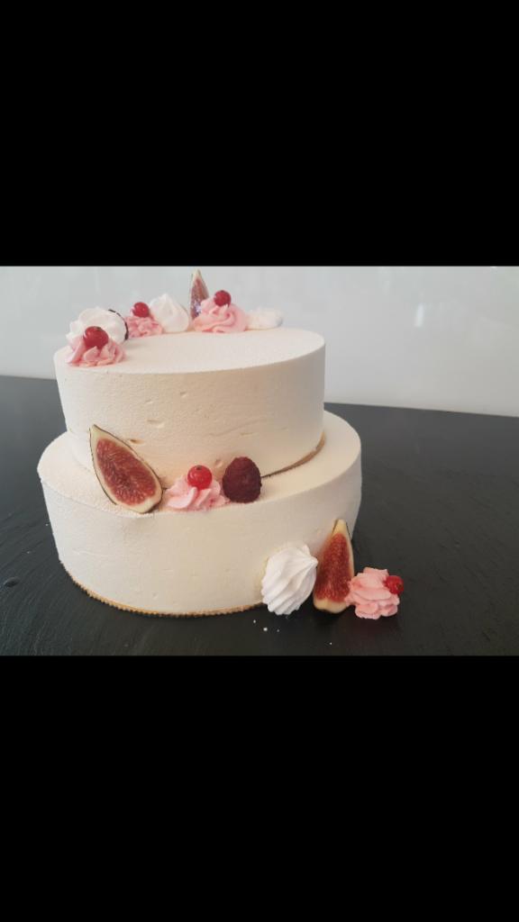 Dwupoziomowy tort z białym kremem, dekorowany figami, malinami, czerwoną porzeczką i bezami na ciemnym blacie.