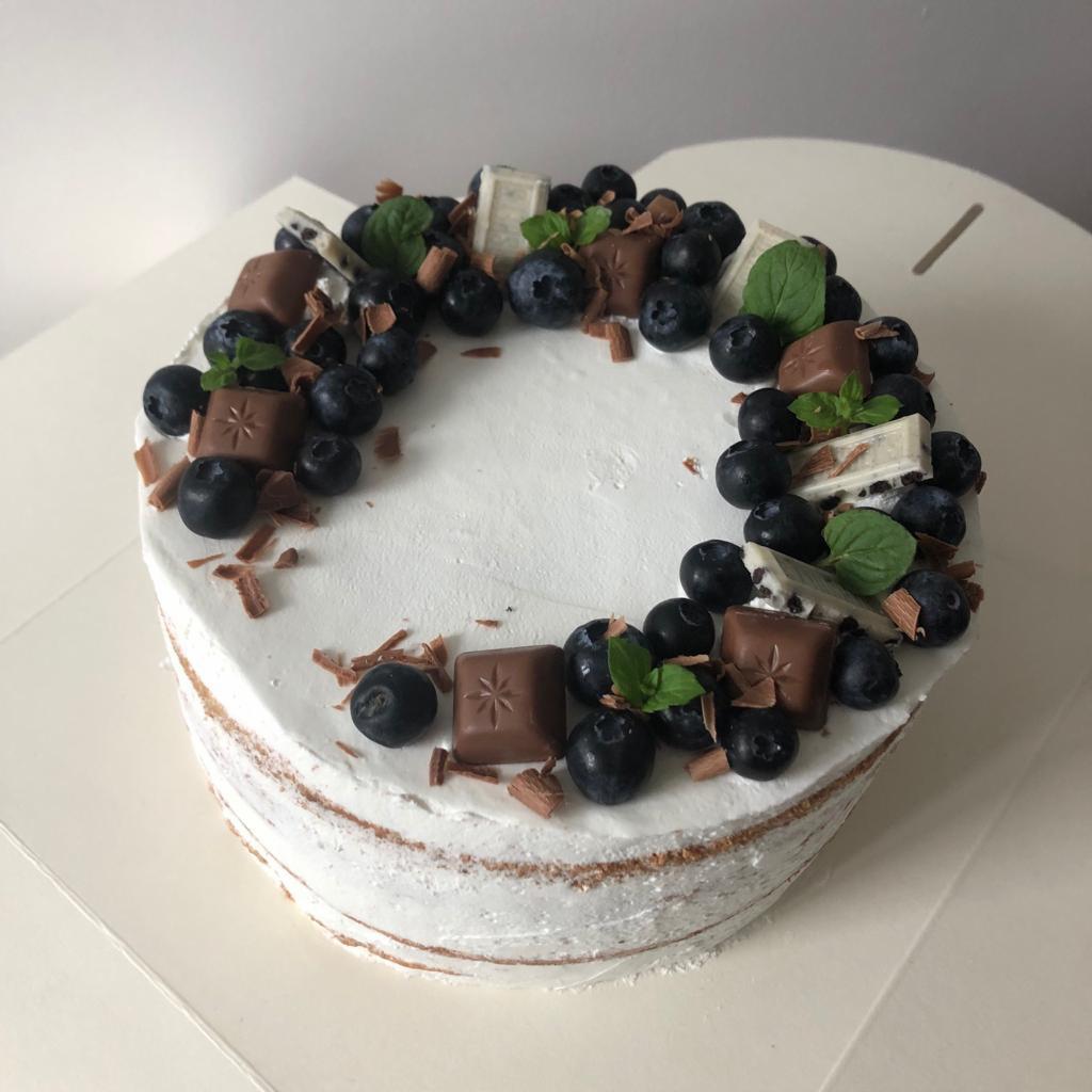 Okrągły tort z białym kremem, udekorowany świeżymi borówkami, kawałkami czekolady mlecznej i białej oraz listkami mięty, tworzącymi wieniec na wierzchu.