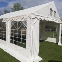 Biały namiot imprezowy z oknami w kształcie łuków, rozstawiony na trawie, z widocznym budynkiem gospodarczym w tle.