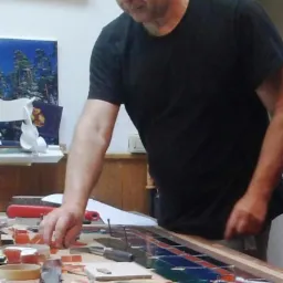 Mężczyzna w okularach dopasowuje elementy witrażu na drewnianym stole warsztatowym, widoczne narzędzia i fragmenty szkła w odcieniach czerwieni, niebieskiego i zieleni.