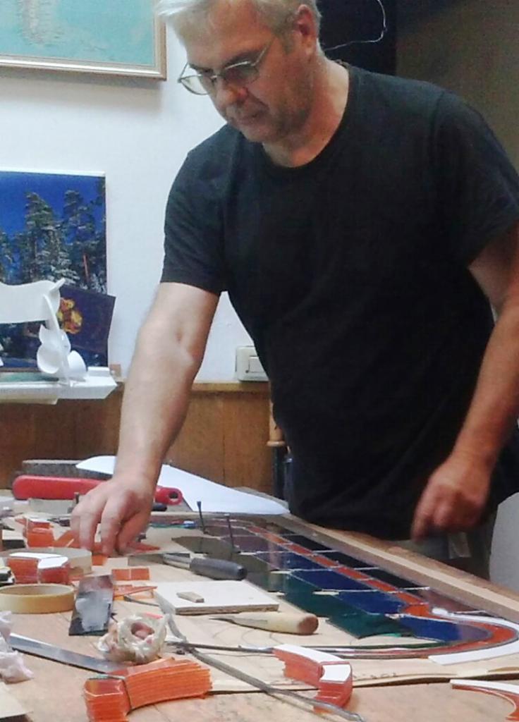 Mężczyzna w okularach dopasowuje elementy witrażu na drewnianym stole warsztatowym, widoczne narzędzia i fragmenty szkła w odcieniach czerwieni, niebieskiego i zieleni.