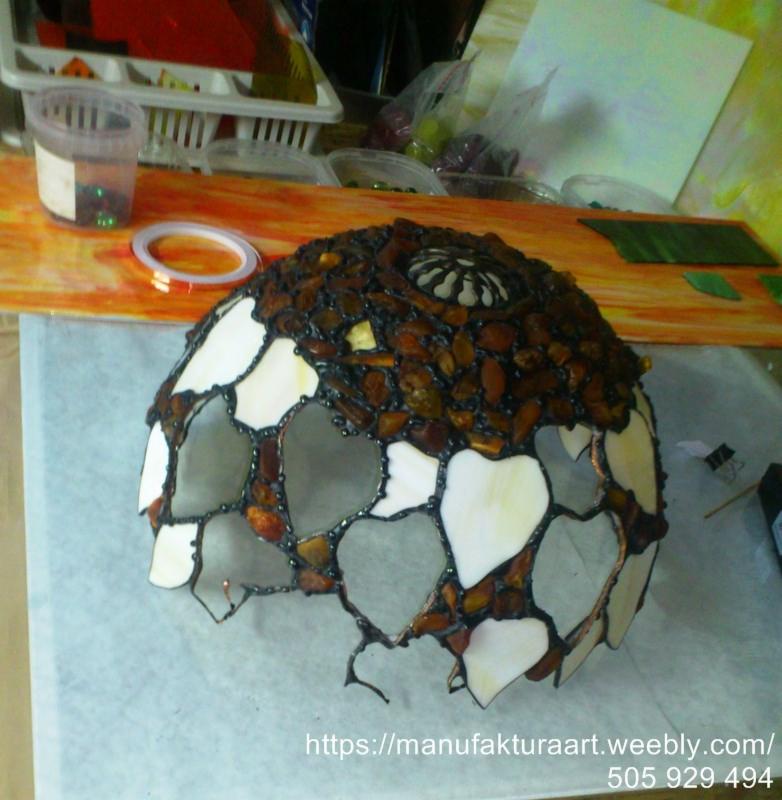 Niedokończony klosz lampy witrażowej w stylu Tiffany, z elementami z brązowego szkła i białymi wstawkami, na stole roboczym z narzędziami i materiałami do witrażu.