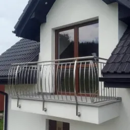 Biały dom z balkonem z chromowaną balustradą na tle ciemnej dachówki i brązowych ram okiennych.