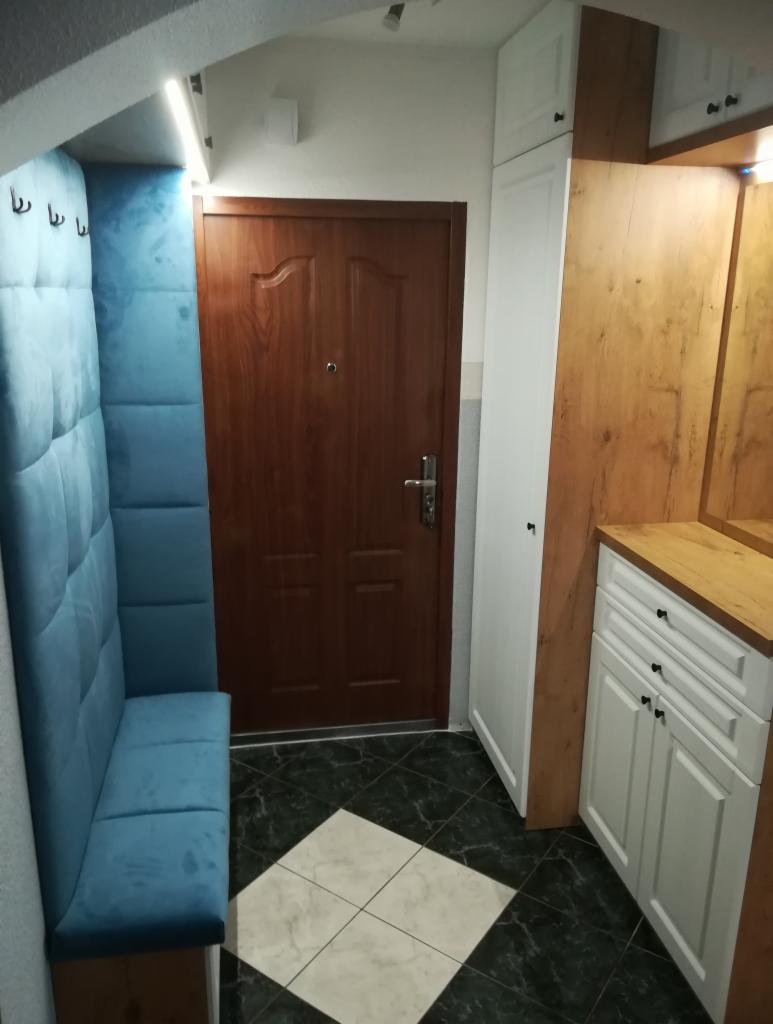 Przedpokój w Ełku z zabudową: tapicerowany, błękitny panel ścienny z siedziskiem, drewniane drzwi wejściowe, szafka z blatem i szafa wnękowa.