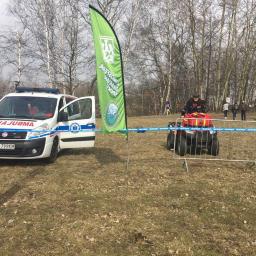 Safety Service - Ambulans i quad ratowniczy na trawiastym terenie, w tle grupa osób i drzewa bez liści, flaga z logo. Otwarta boczna szyba ambulansu.