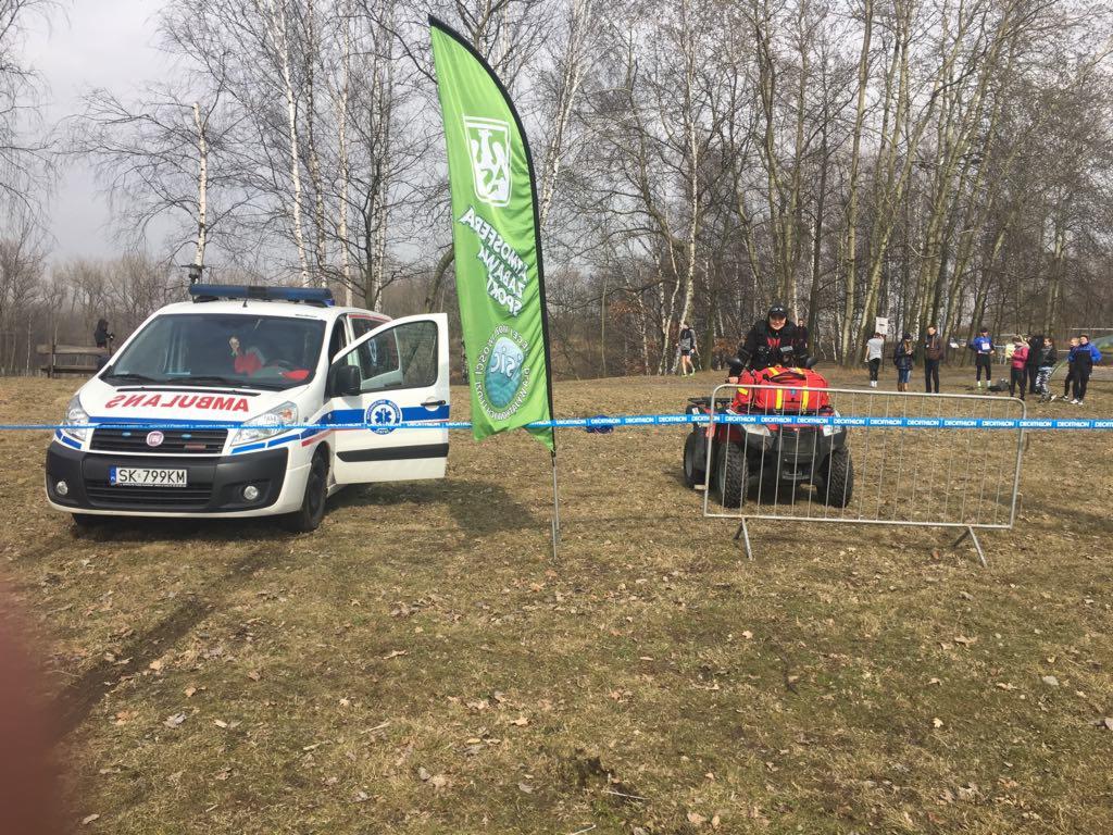 Ambulans i quad ratowniczy na trawiastym terenie, w tle grupa osób i drzewa bez liści, flaga z logo. Otwarta boczna szyba ambulansu.