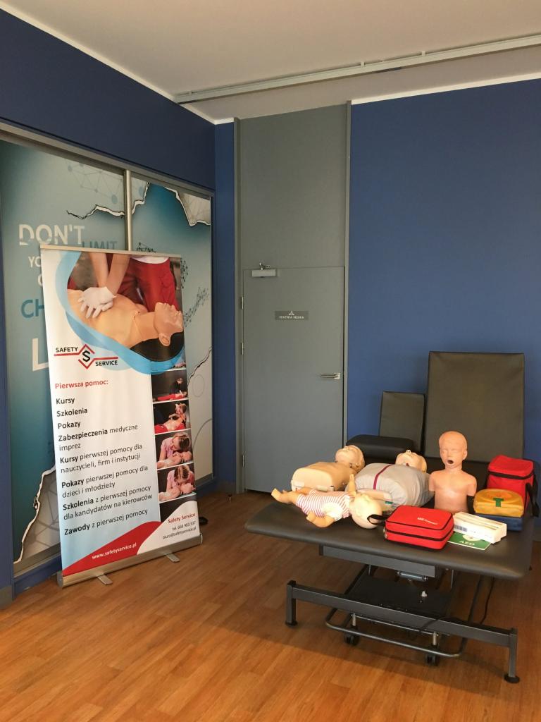 Sala szkoleniowa z manekinami do nauki pierwszej pomocy na stole, obok baner reklamowy z ofertą kursów i szkoleń z zakresu pierwszej pomocy.