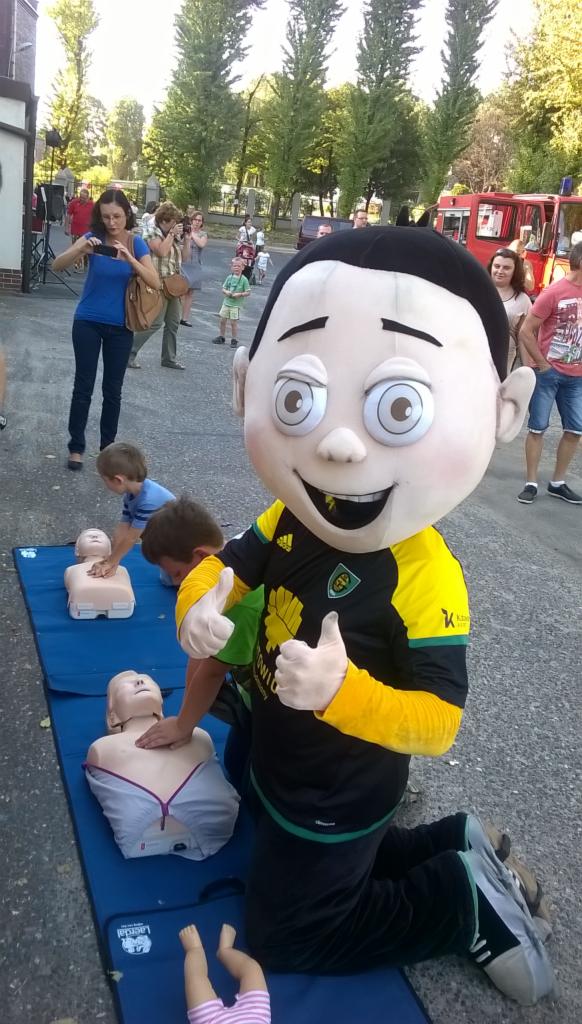 Mascot gives thumbs up as children practice CPR on training dummies during an outdoor first aid demonstration, with onlookers and a red fire truck in the background.