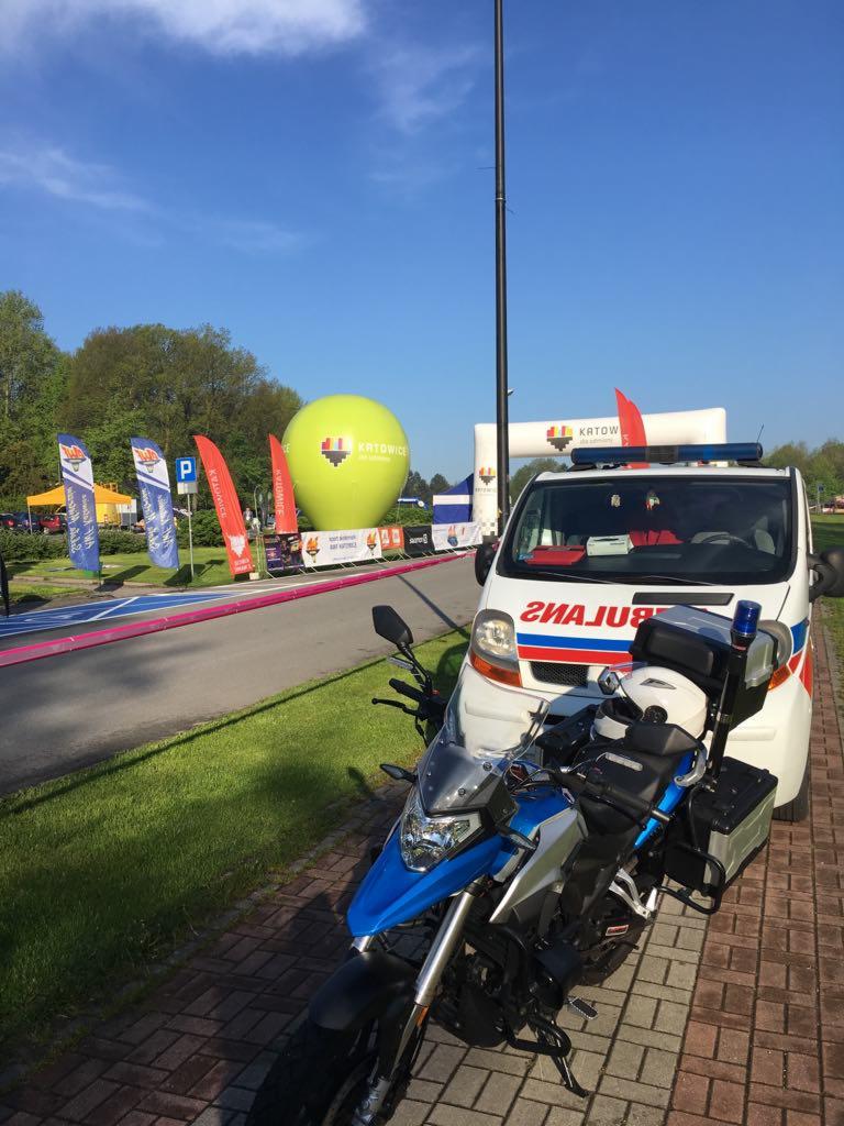 Motocykl ratunkowy z niebieskim błyskowym światłem i ambulans na tle zielonego balonu z napisem Katowice, flaga i brama startowa, błękitne niebo