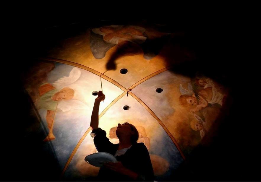 Praca konserwatorska: osoba z paletą i pędzlem odnawia malowidło sufitowe z aniołami w kościele, oświetlone punktowym światłem.