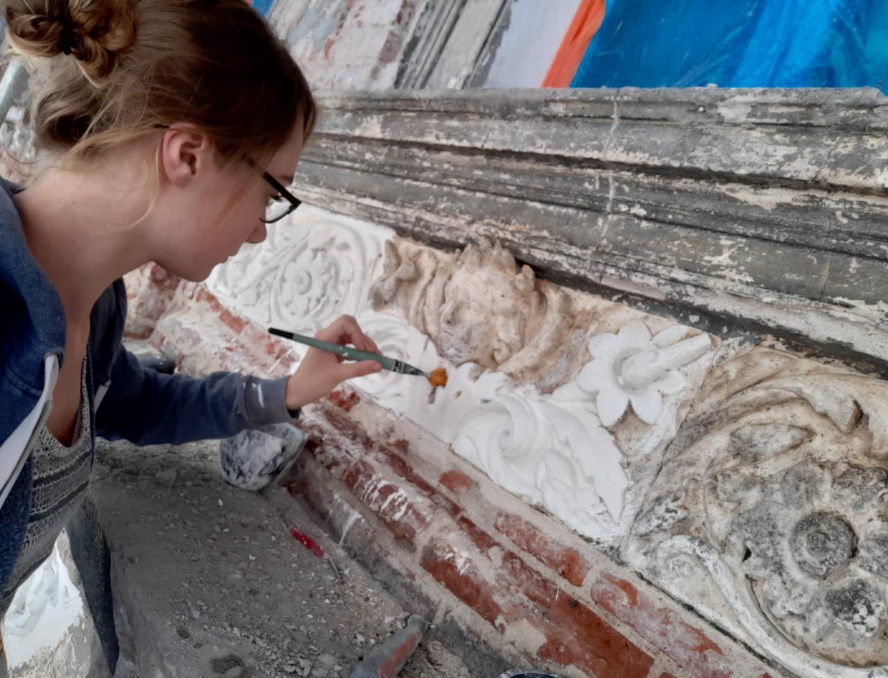 Kobieta w okularach delikatnie odnawia ozdobny, kamienny element architektoniczny na ceglanej ścianie za pomocą pędzla, uwydatniając detale wzoru.