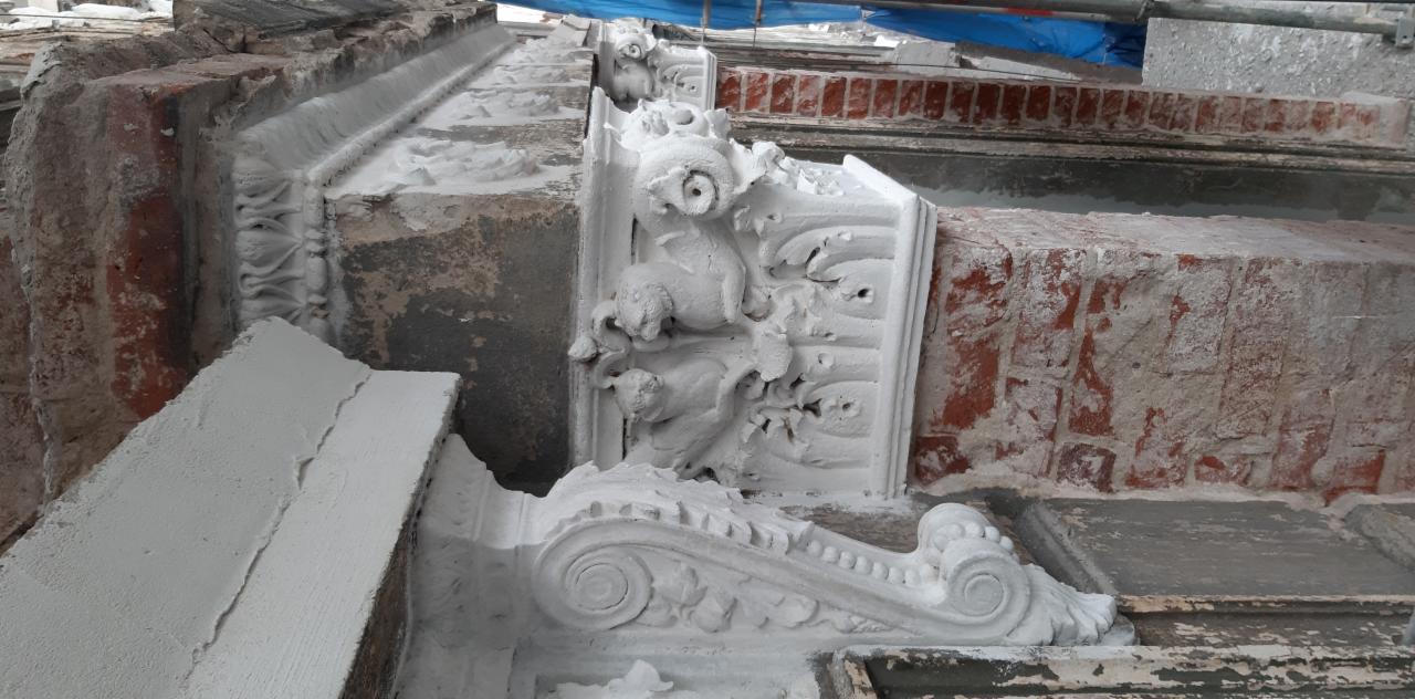 Fragment elewacji budynku z dekoracyjnymi elementami architektonicznymi, w trakcie prac renowacyjnych, pokryty warstwą białego tynku lub farby. Widoczne detale rzeźbiarskie przedstawiające...