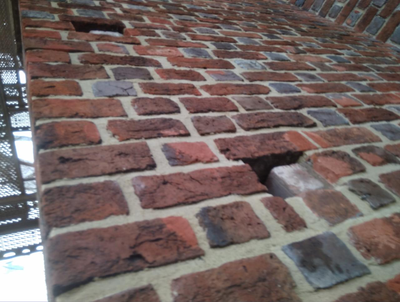 Fragment ściany z czerwonej cegły z widocznymi ubytkami i nierównościami, sfotografowany pod kątem z perspektywy żabiej, z fragmentem metalowej konstrukcji po lewej stronie.