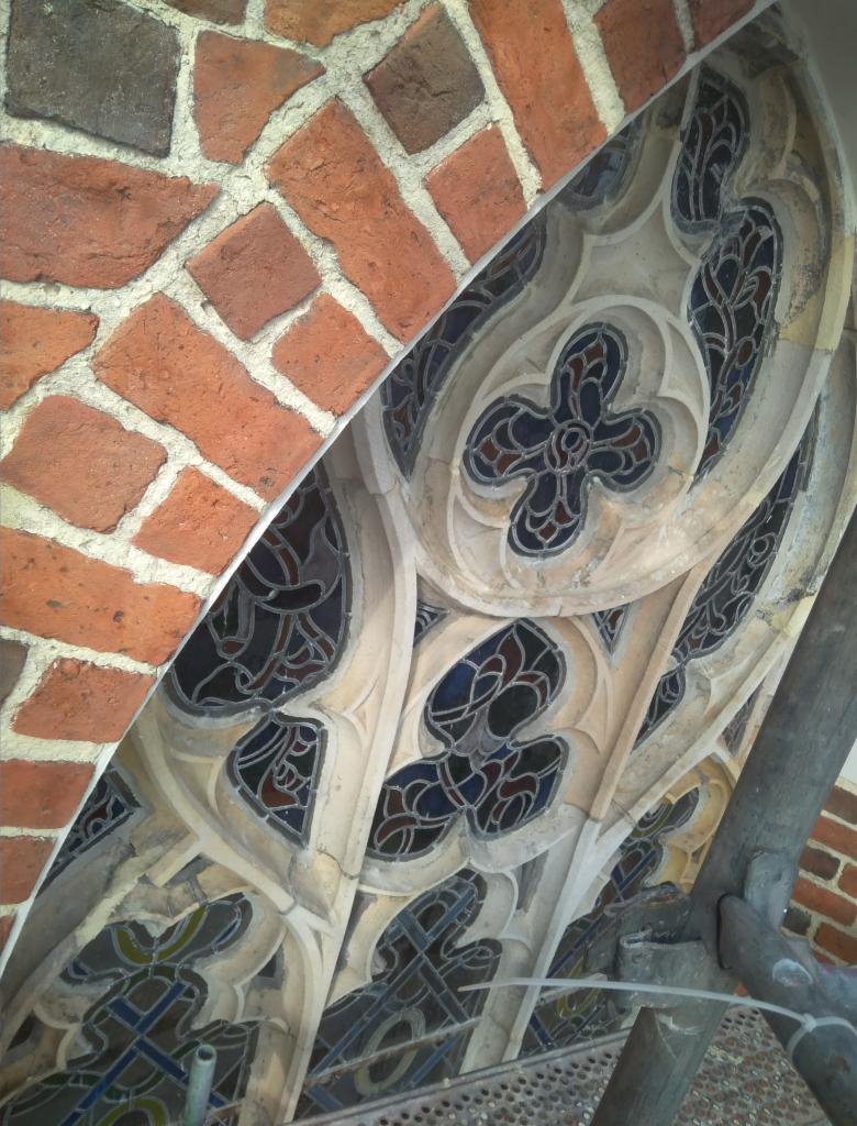 Fragment zabytkowej elewacji z cegły i kamiennych zdobień okiennych z witrażami, widoczne ślady renowacji i rusztowanie.