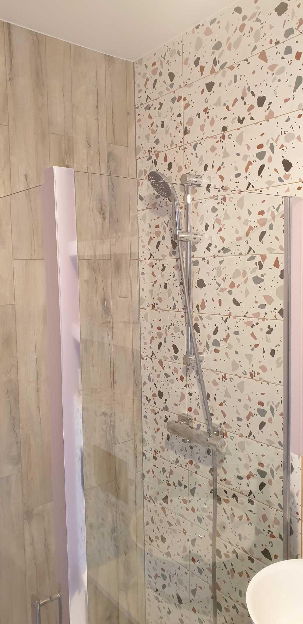 Nowoczesna kabina prysznicowa z chromowaną armaturą na tle ściany wyłożonej płytkami imitującymi lastryko oraz drewnopodobnymi płytkami na sąsiedniej ścianie.