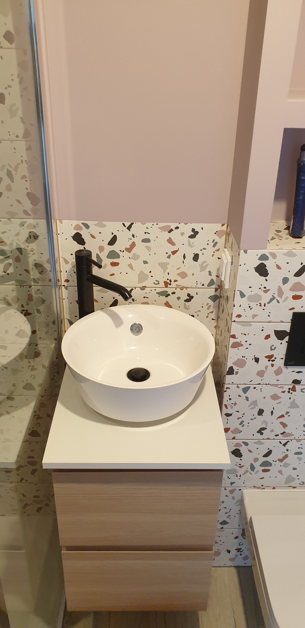 Nowoczesna łazienka z okrągłą białą umywalką nablatową, czarną armaturą i terazzo płytkami na ścianie oraz szafką pod umywalką z drewnopodobnym frontem.