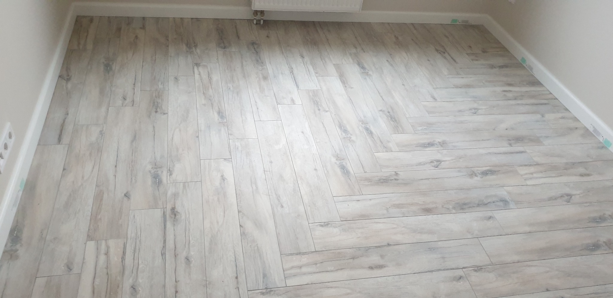 Ułożona pod kątem jasna podłoga z płytek imitujących drewno w pomieszczeniu w Kamieniu Pomorskim, widoczne listwy przypodłogowe i fragment grzejnika.