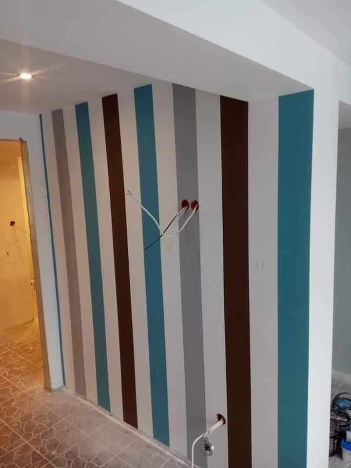 Ściana w pionowe pasy w odcieniach błękitu, szarości, brązu i bieli, z widocznymi przewodami elektrycznymi wystającymi ze ściany, podłoga częściowo zakryta folią malarską, widoczne puszki z farbą...