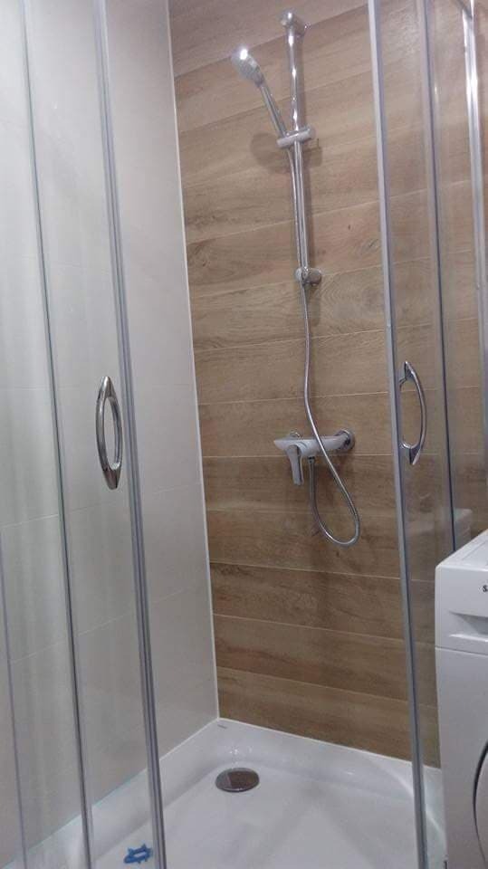 Kabiną prysznicowa z chromowaną armaturą na tle ściany wyłożonej płytkami imitującymi drewno, widoczna częściowo pralka.