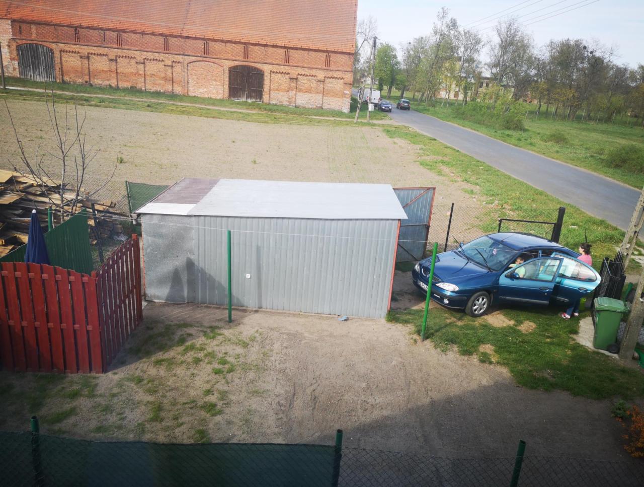 Blaszany garaż z otwartymi drzwiami, obok zaparkowany niebieski samochód, w tle budynek gospodarczy z czerwonej cegły i droga.