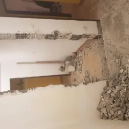 Wyburzony fragment ściany działowej, odsłonięta konstrukcja i gruz na podłodze, widoczne przejście do kolejnego pomieszczenia.