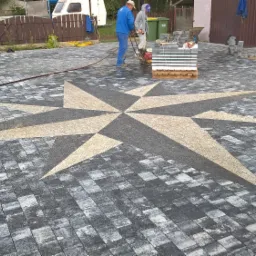 Ułożona nawierzchnia z kostki brukowej z wzorem gwiazdy w centralnej części, dwaj pracownicy używają zagęszczarki w tle, paleta z kostką obok.