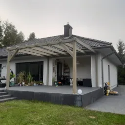 Nowoczesny dom jednorodzinny z szarą drewnianą pergolą nad tarasem, widok z trawnika, w tle drzewa iglaste.