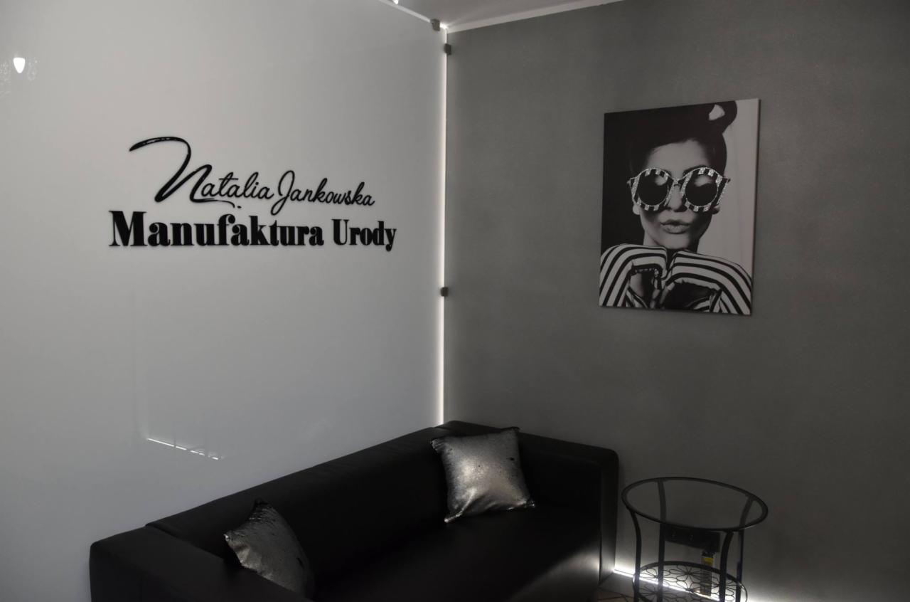 Poczekalnia salonu kosmetycznego: czarna sofa z połyskującymi poduszkami, szklana ściana z logo 'Natalia Jankowska Manufaktura Urody' i grafika kobiety w okularach.