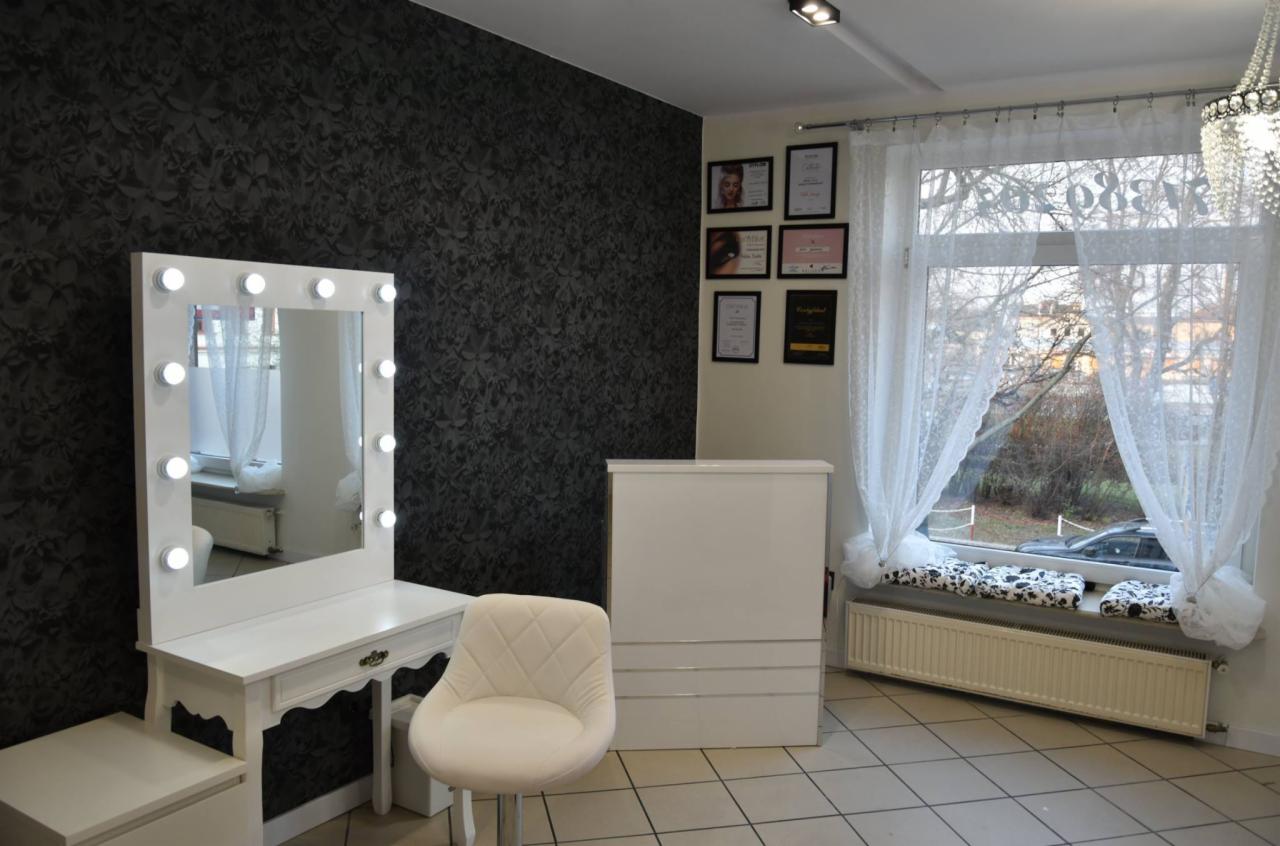 Wnętrze jasnego studia w kremowych odcieniach z toaletką z lustrem otoczonym żarówkami, białym krzesłem i recepcją na tle ściany w ciemne kwiatowe wzory oraz okna z białymi firankami.