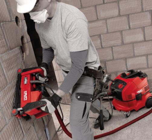 Pracownik w masce przeciwpyłowej wycina bruzdy w ścianie z cegły za pomocą frezarki Hilti DC-SE20 podłączonej do odkurzacza przemysłowego, widoczne kable i narzędzia na pasie.