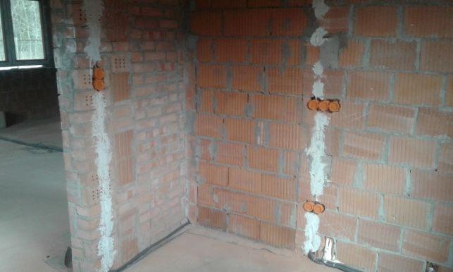 Murowana ściana z bruzdami wypełnionymi gipsem, w których osadzone są pomarańczowe puszki elektryczne, widok na surowy stan wykończenia wnętrza.