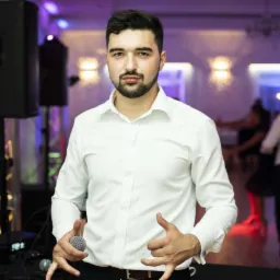 Mężczyzna w białej koszuli trzyma mikrofon, na drugim planie sprzęt nagłośnieniowy i tańczący ludzie w sali balowej z oświetleniem w odcieniach fioletu.