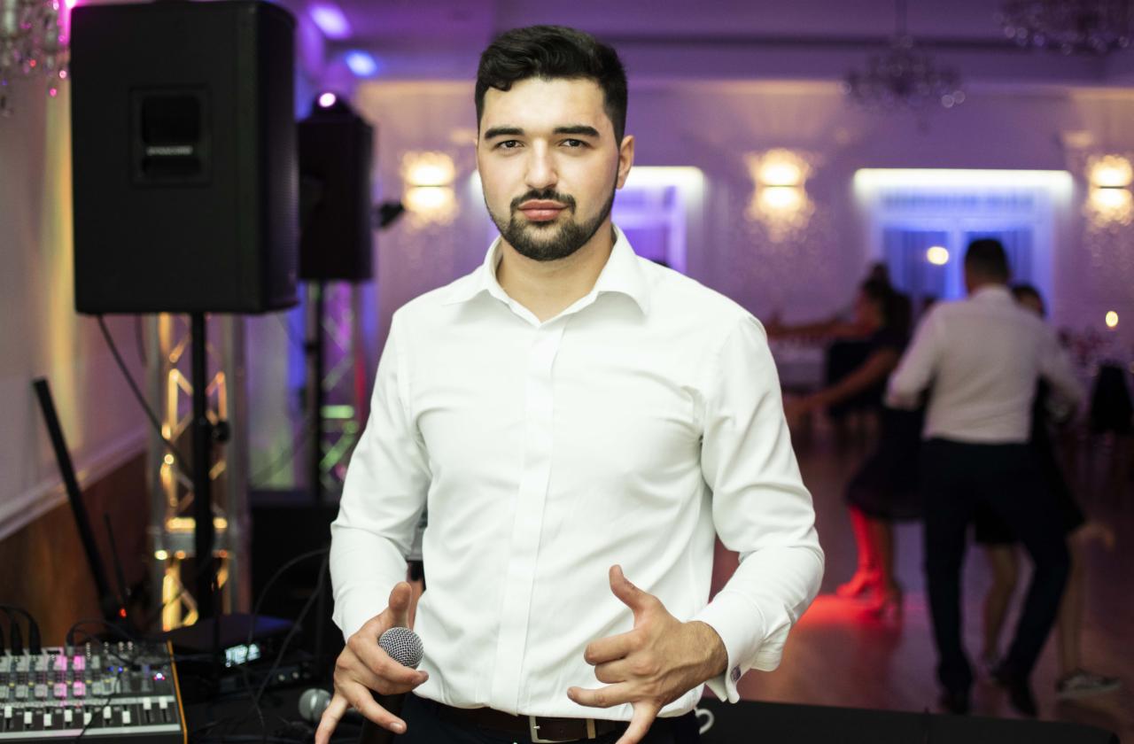 Mężczyzna w białej koszuli trzyma mikrofon, na drugim planie sprzęt nagłośnieniowy i tańczący ludzie w sali balowej z oświetleniem w odcieniach fioletu.