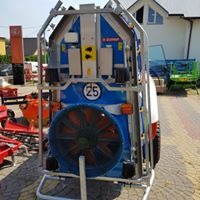 Niebiesko-biała maszyna rolnicza z wentylatorem w metalowej ramie, widoczna z tyłu na tle brukowanej powierzchni i budynków.