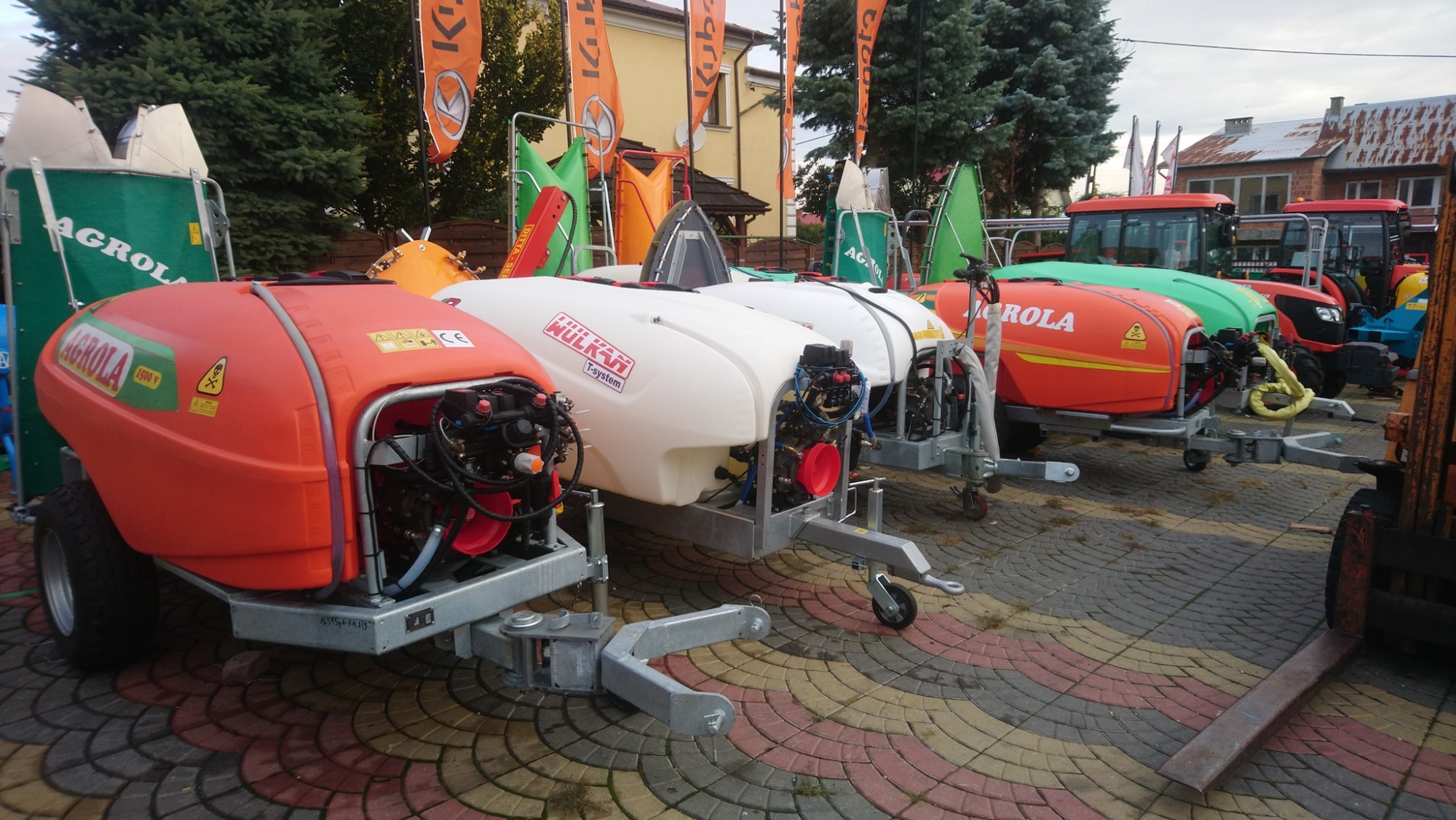 Rząd opryskiwaczy polowych Agrola i Wulkan T-system na przyczepach, zaparkowanych na brukowanym placu, z widocznymi flagami reklamowymi w tle.