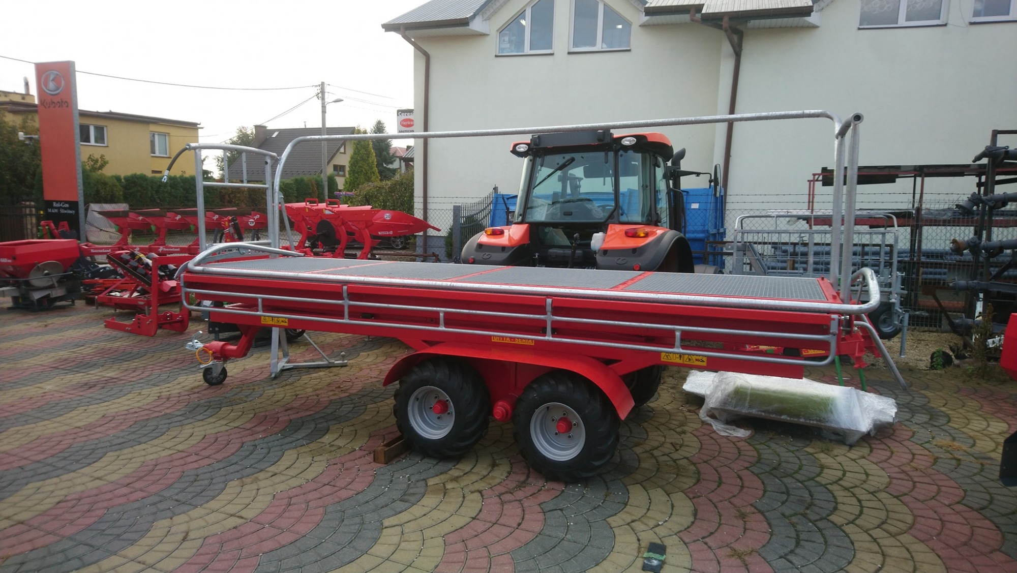 Czerwona przyczepa rolnicza z platformą i relingami, widoczna na tle ciągnika w Sandomierzu, prezentowana na brukowanym placu.