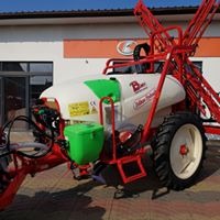 Czerwono-biały opryskiwacz polowy z zielonym zbiornikiem dodatkowym, stojący na placu przed budynkiem z pomarańczowym akcentem.