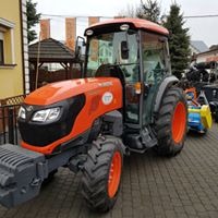 Pomarańczowy ciągnik rolniczy Kubota M5071 N z kabiną, zaparkowany na brukowanym podwórku, z widocznymi elementami osprzętu rolniczego w tle.