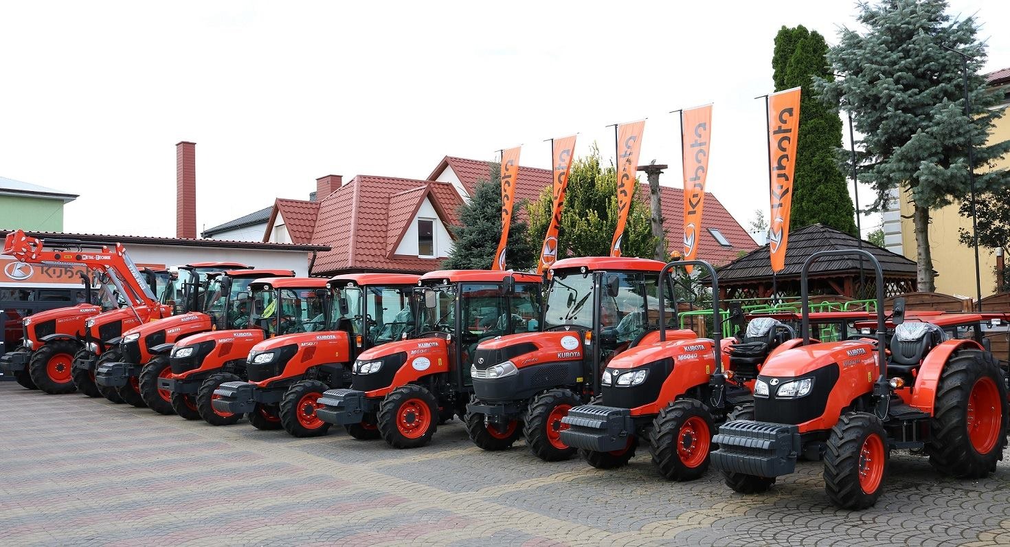 Rząd nowych, pomarańczowych traktorów Kubota ustawionych na placu przed budynkiem z czerwoną dachówką, z widocznymi flagami firmowymi.