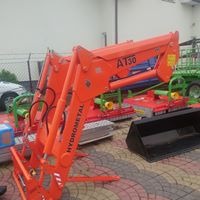 Pomarańczowy osprzęt rolniczy Hydrometal AT30, prawdopodobnie ładowacz czołowy, na tle innych maszyn rolniczych i zaparkowanego samochodu.