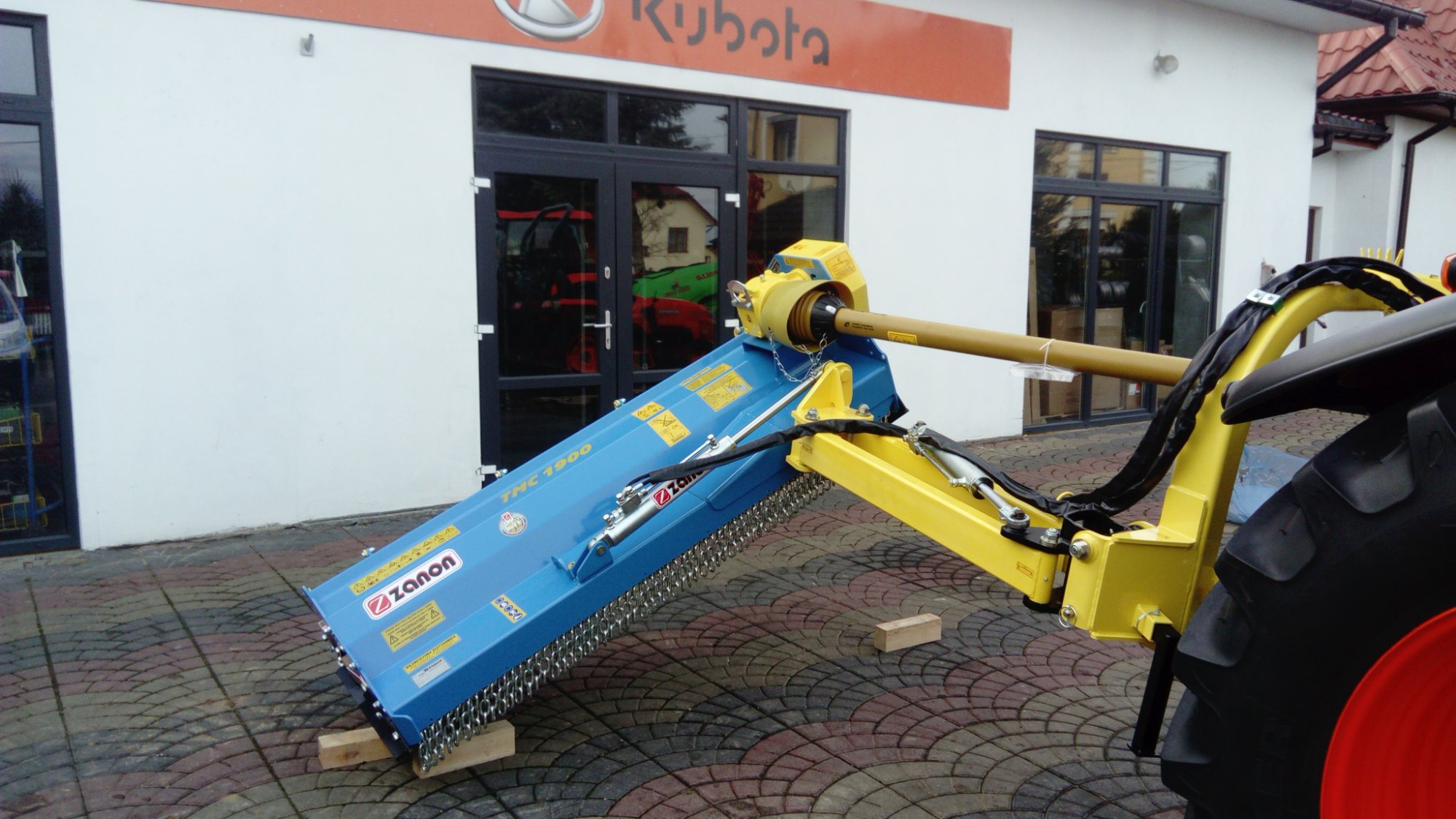 Niebieska kosiarka bijakowa Zanon TMC 1900 podłączona do ciągnika Kubota, stojąca na drewnianych klockach przed salonem sprzedaży.