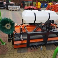 Pomarańczowe urządzenie czyszczące z białą plastikową beczką na płyn, zieloną szczotką i czarną metalową ramą, stojące na kostce brukowej. W tle widoczny plac z maszynami budowlanymi.